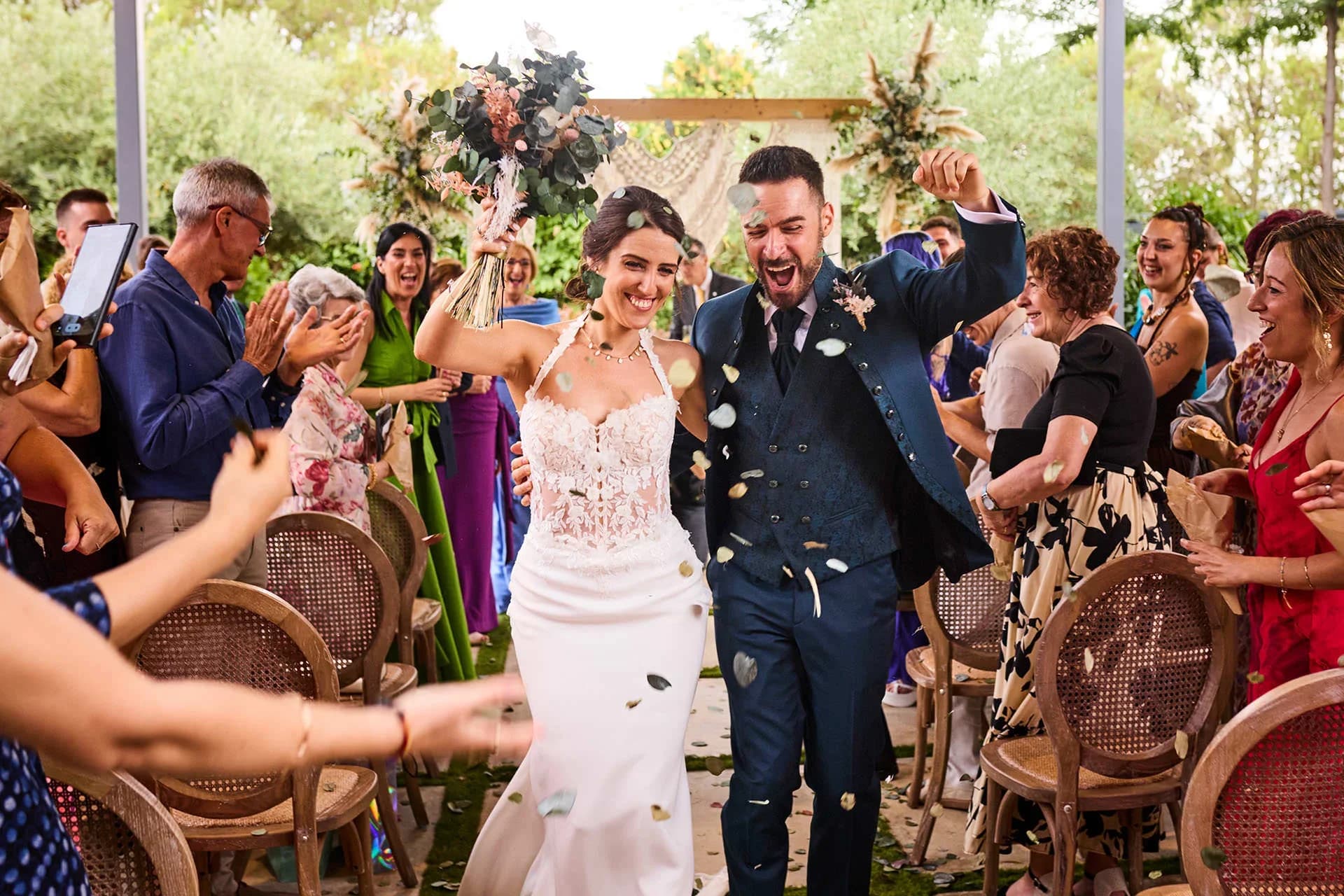 Los recién casados caminan sonrientes entre los invitados que aplauden y lanzan pétalos, ella con el ramo en alto y él celebrando con el puño cerrado justo después de la ceremonia.
