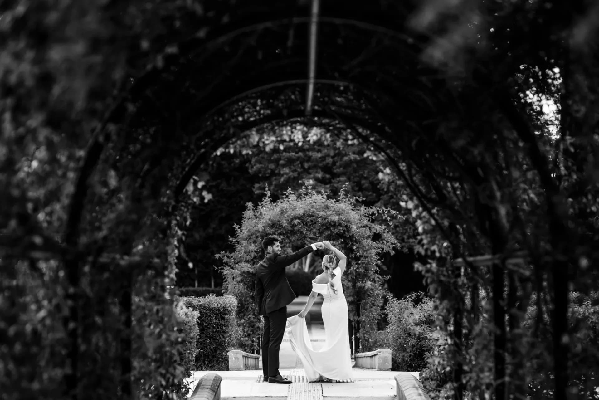 Retrato en blanco y negro de los novios bailando bajo un túnel de pérgola cubierto de vegetación, él la hace girar cogida de la mano mientras ella sonríe con el vestido en movimiento.