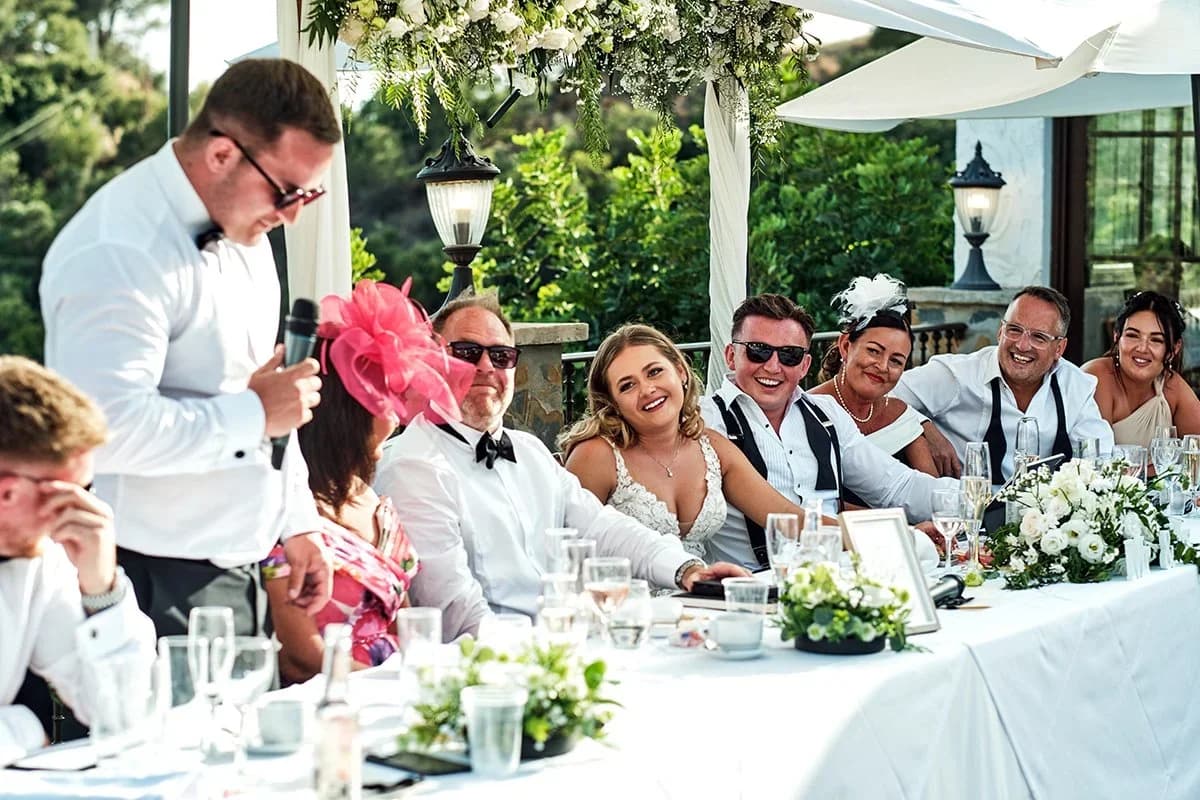 Mesa presidencial en una terraza al aire libre durante un banquete de boda, un hombre de pie con micrófono da un discurso mientras la novia y otros invitados, algunos con gafas de sol y tocados, ríen a carcajadas bajo un arco floral.