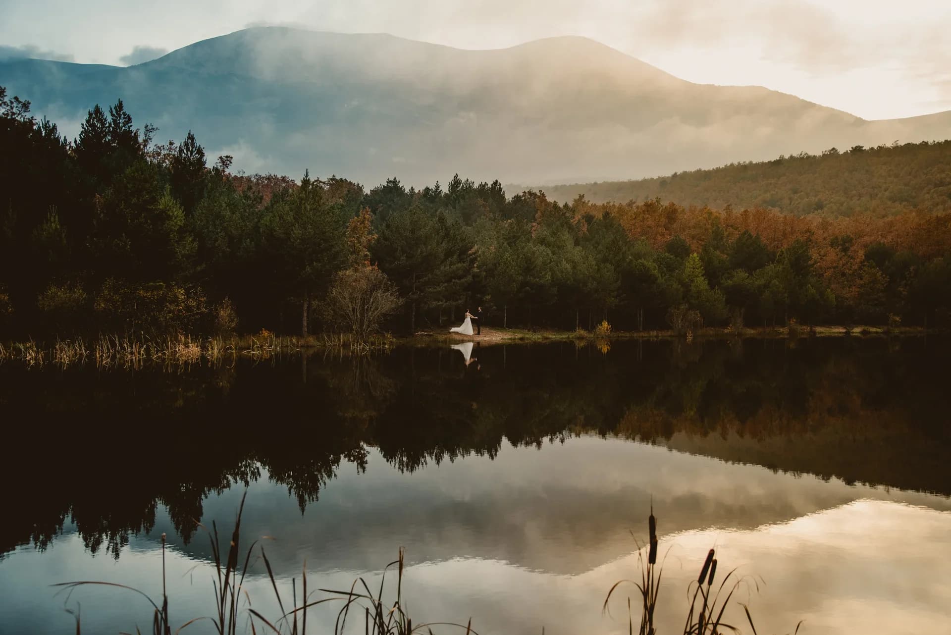 Paisaje de montaña con niebla y un lago en calma que refleja con nitidez el bosque otoñal y las cumbres, con la pareja de novios como pequeñas figuras junto a la orilla.