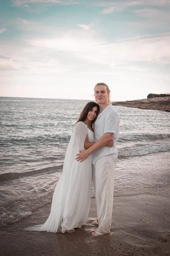 Pareja abrazada de pie en la orilla del mar, vestidos de blanco, mirando a cámara con el horizonte marino y una cala rocosa al fondo, cielo nublado suave.
