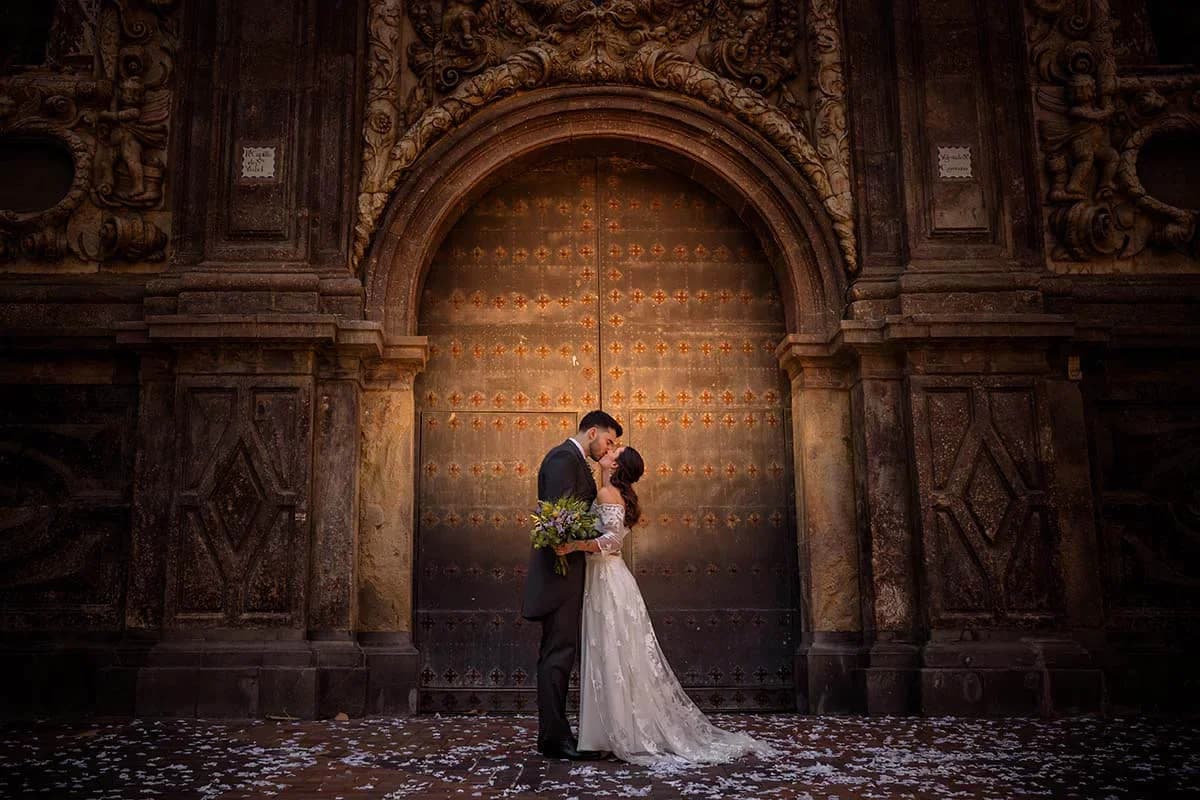Los novios se besan de pie ante una enorme puerta de madera claveteada y tallada de una catedral, iluminados con luz cálida y rodeados de confeti blanco esparcido por el suelo.