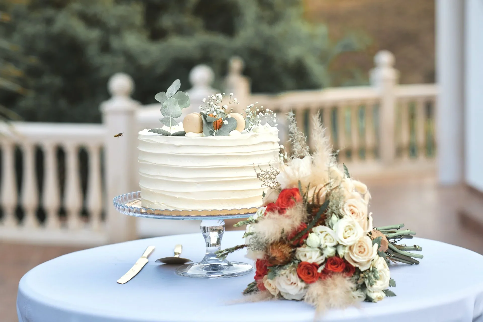 Detalle de una tarta nupcial de un piso decorada con macarons, ramitas de eucalipto y flores blancas junto a un ramo de novia de rosas rojas y blancas con pampas, sobre una mesa con mantel azul claro y balaustrada desenfocada al fondo.