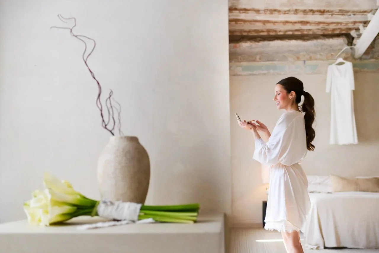 La novia, en bata blanca, se retoca el maquillaje mirándose en un espejo de mano dentro de una habitación luminosa de estilo rústico-mediterráneo, con el vestido colgado al fondo y un ramo de calas sobre una mesa en primer plano.