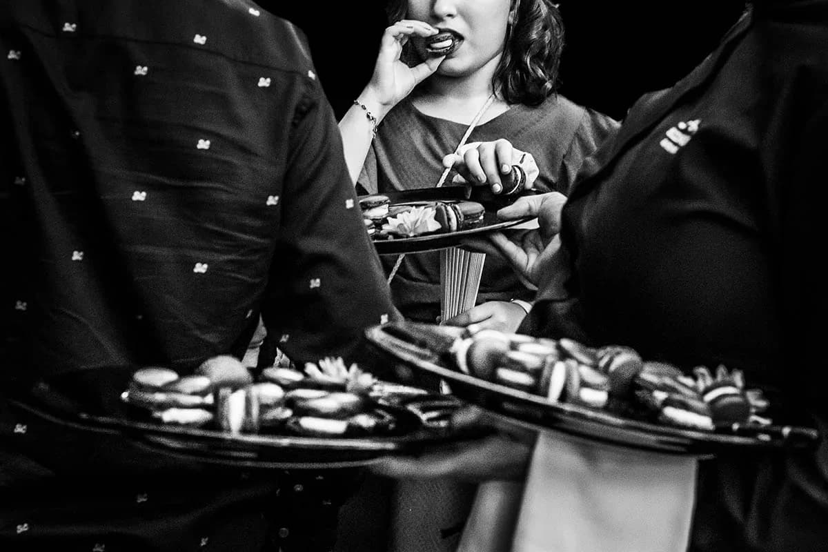 En blanco y negro y con fuerte desenfoque, una invitada se lleva un macaron a la boca mientras dos camareros sostienen bandejas repletas de macarons y pastelitos durante el cóctel.