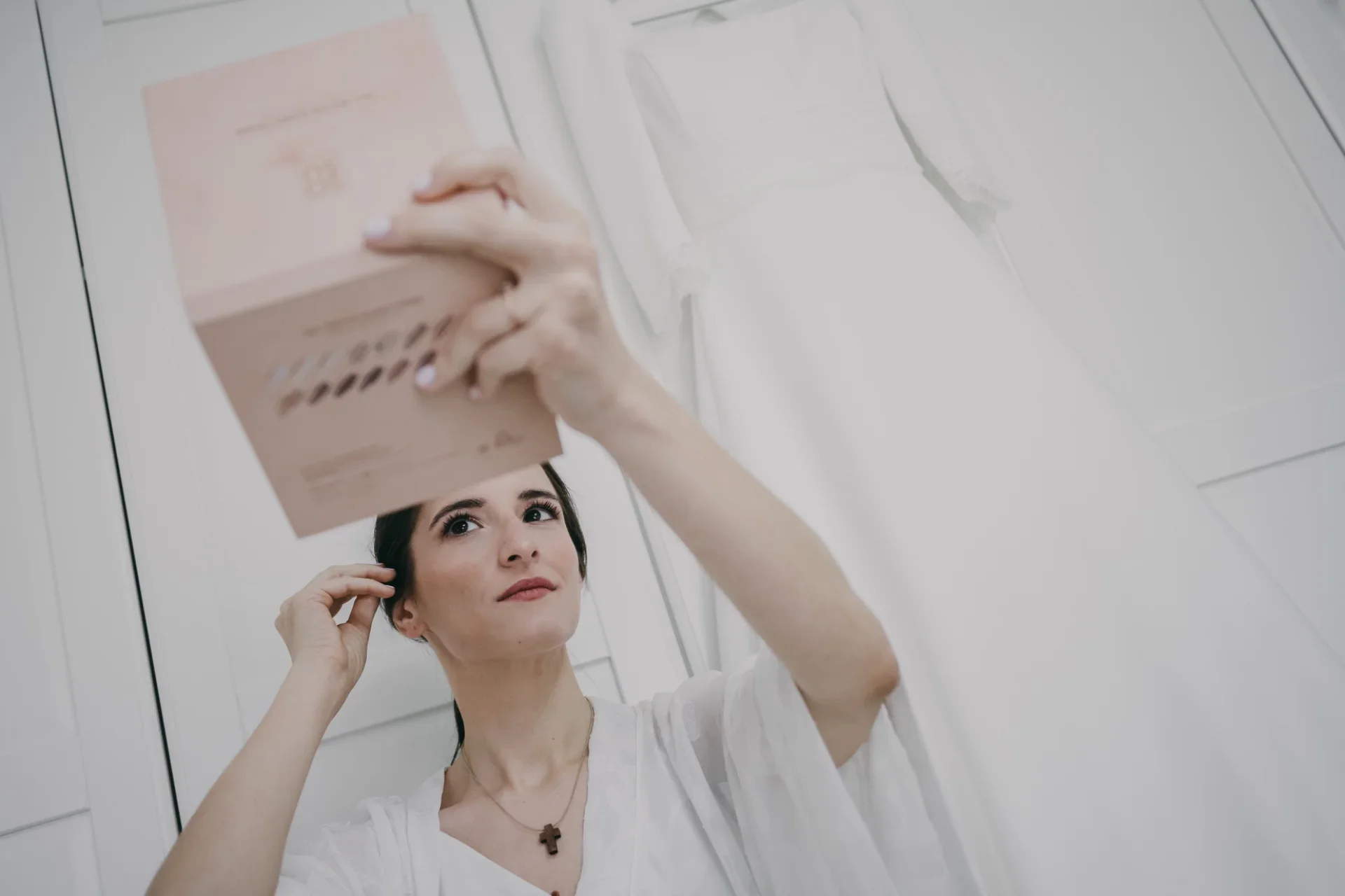 La novia, en bata blanca, sostiene sobre su cabeza una caja rosa de invitaciones o regalo y la observa con atención, en una habitación luminosa y sobreexpuesta con el vestido de novia colgado desenfocado al fondo.