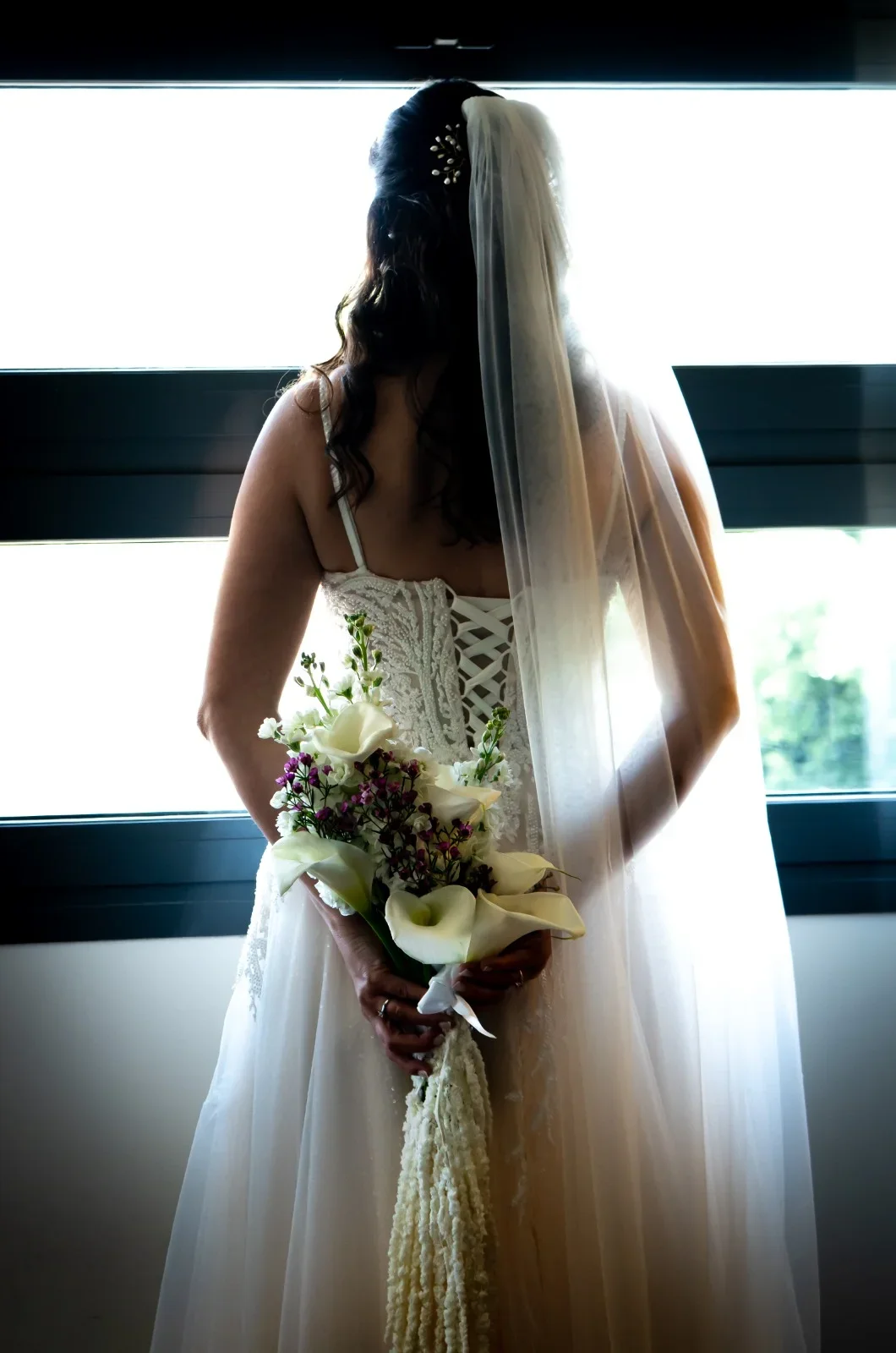 La novia de espaldas frente a un gran ventanal a contraluz, sujeta tras la espalda un ramo de calas blancas y flores moradas, con el velo y el corsé del vestido en primer plano.
