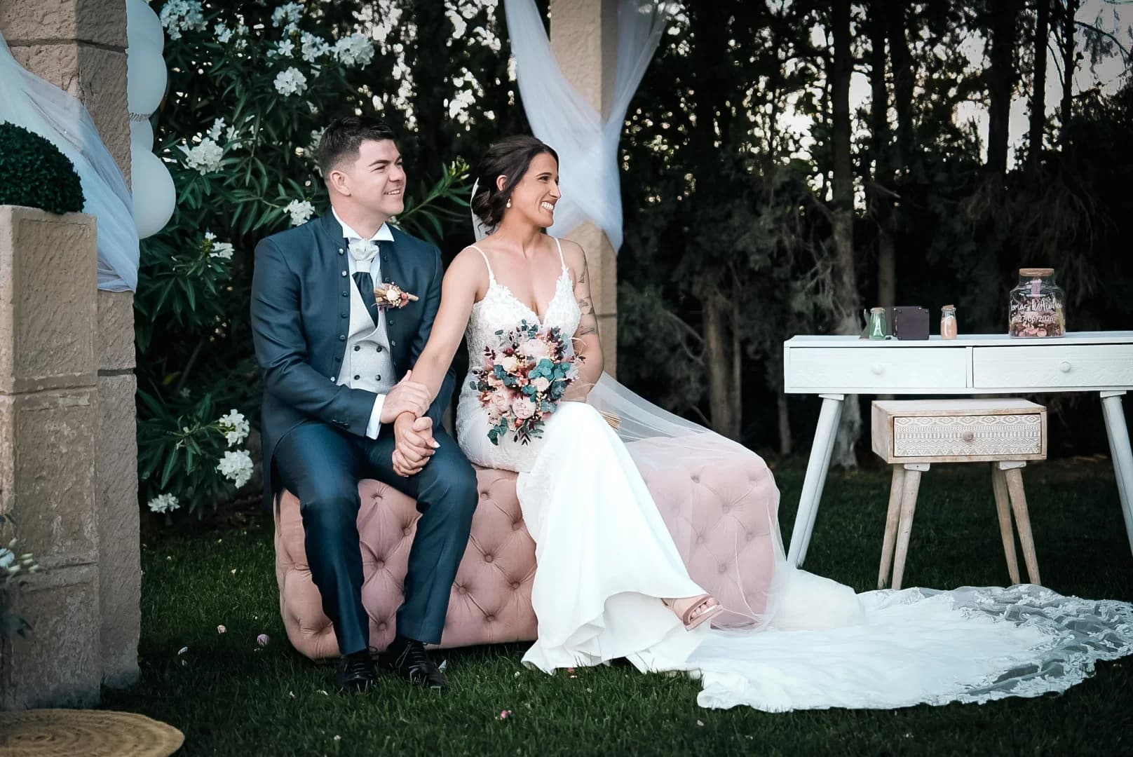 Los novios sentados juntos y sonrientes en un banco tapizado rosa bajo un cenador de piedra con telas blancas, cogidos de la mano, con un jardín de pinos al fondo en la hora dorada.