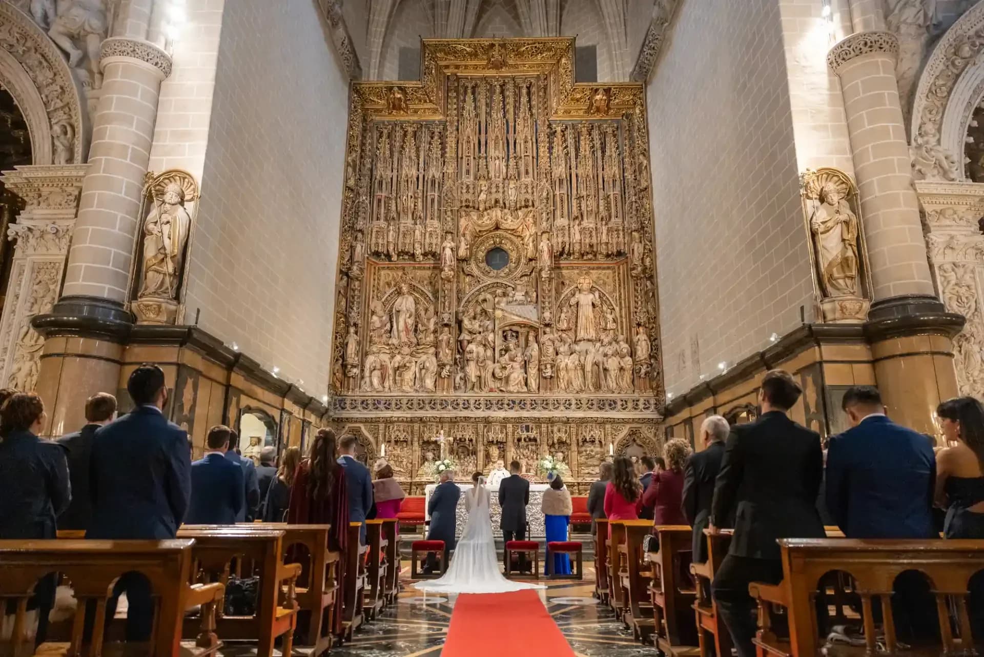 Vista general del interior de una iglesia gótica durante la ceremonia, con los novios ante un retablo dorado monumental, alfombra roja en el pasillo central y los invitados de pie en los bancos.