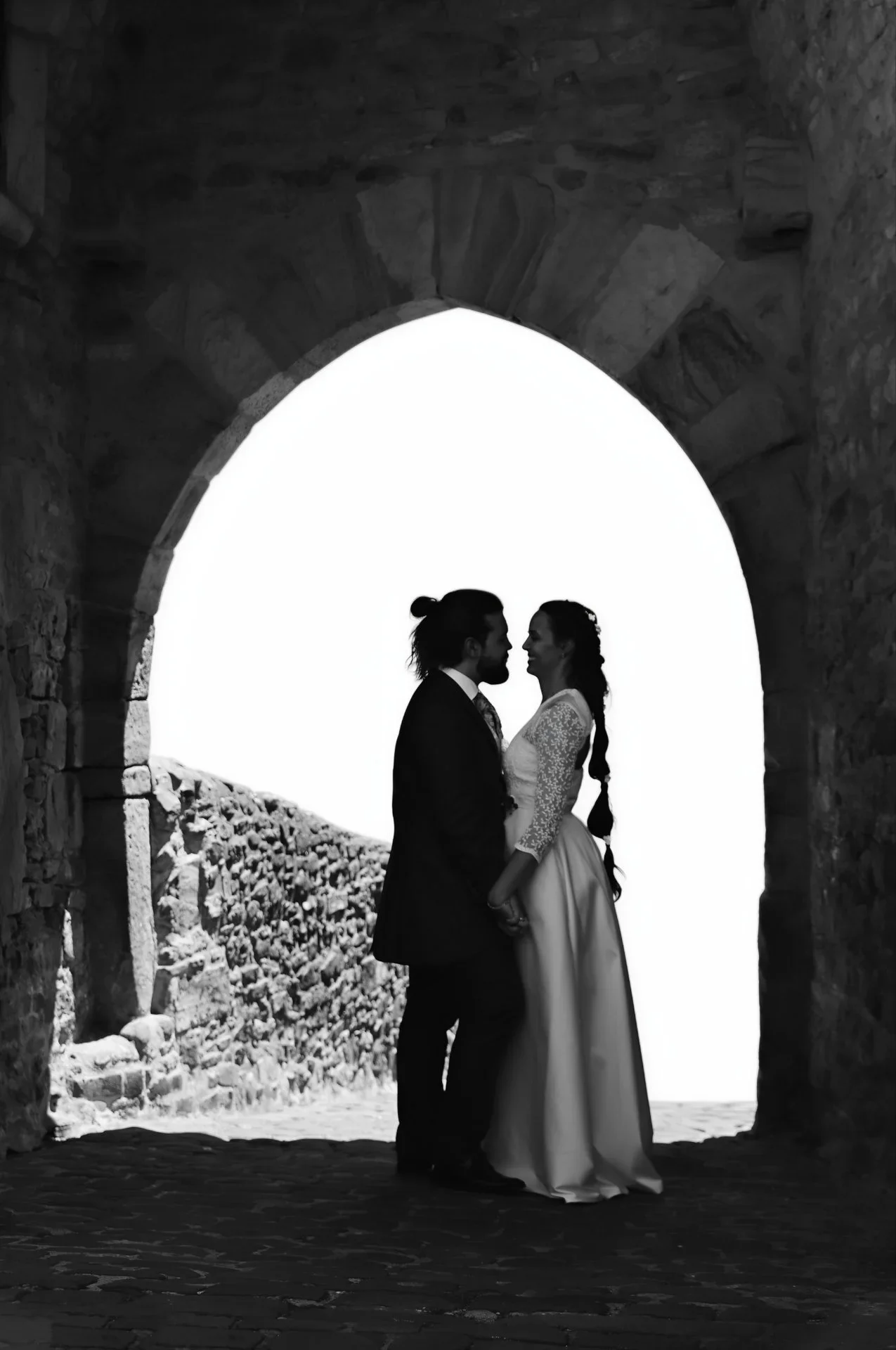 Silueta en blanco y negro de la pareja a punto de besarse bajo un arco de piedra en ruinas, recortada contra un cielo blanco totalmente sobreexpuesto que crea un fuerte contraste dramático.