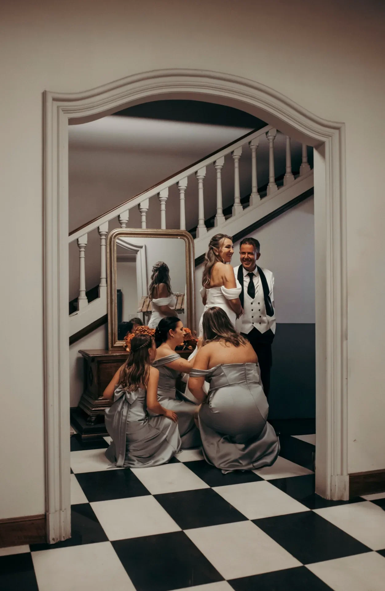 Escena de preparativos captada a través de un arco de puerta, la novia sonríe junto a su padre mientras tres damas de honor con vestidos grises arrodilladas ajustan la cola del vestido, con escalera y espejo dorado de fondo sobre suelo ajedrezado.