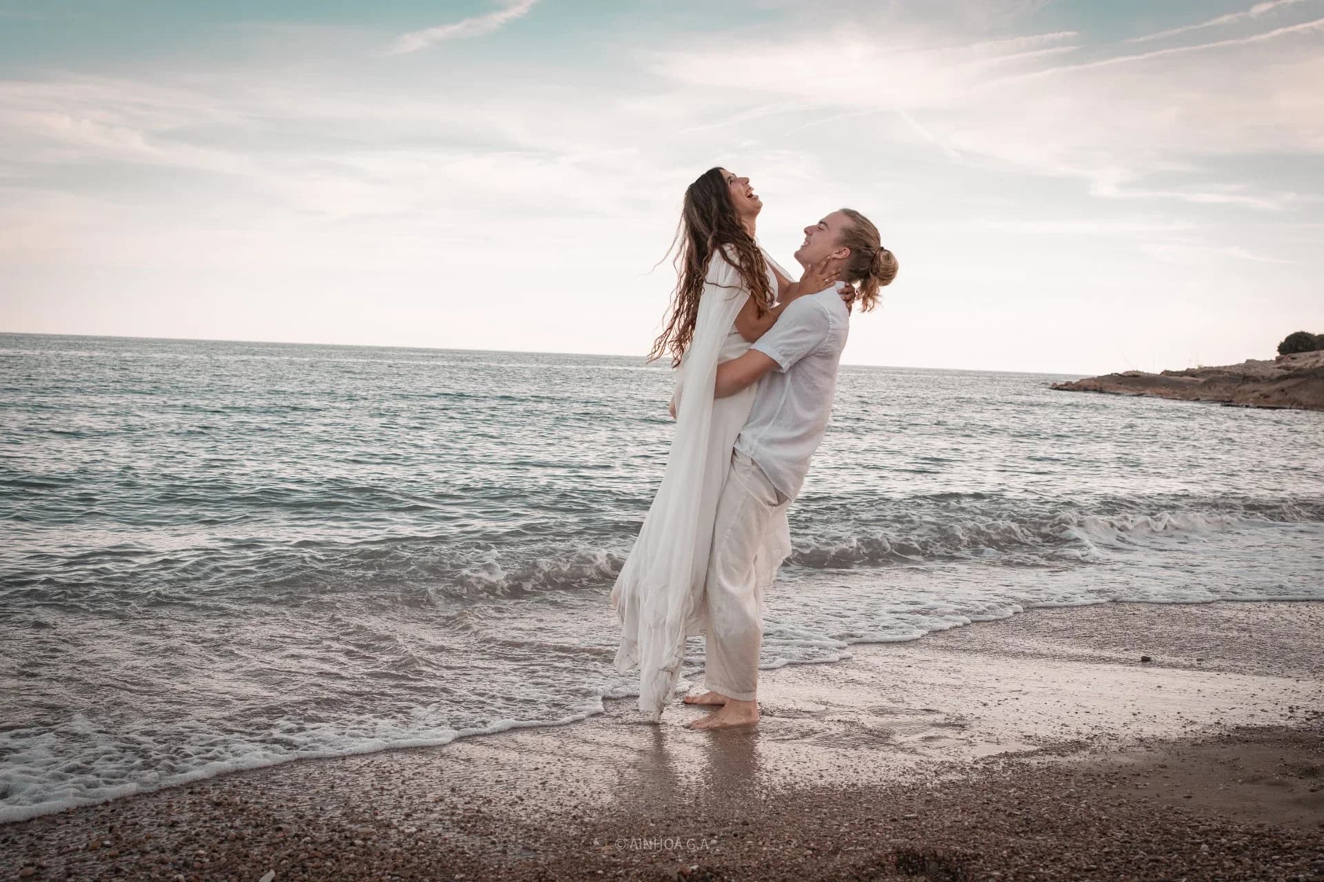 El chico sostiene en volandas a su pareja entre la orilla del mar, ambos riendo mirándose hacia arriba, con el mar y el cielo del atardecer de fondo.