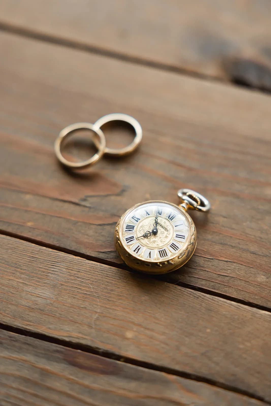 Fotografía de detalle sobre una mesa de madera rústica que muestra dos alianzas doradas junto a un reloj de bolsillo antiguo con numeración romana, evocando el paso del tiempo y la tradición.