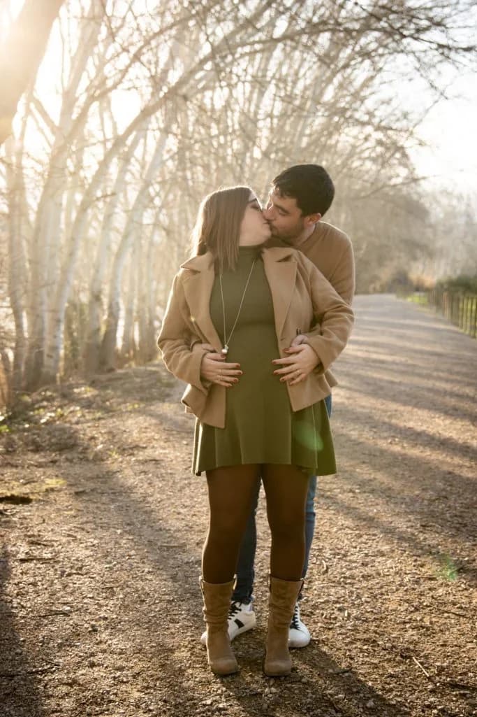 Pareja besándose de pie en un camino de tierra bordeado de árboles sin hojas, a contraluz invernal, ella lleva abrigo camel y vestido verde, él abrigo camel y vaqueros.