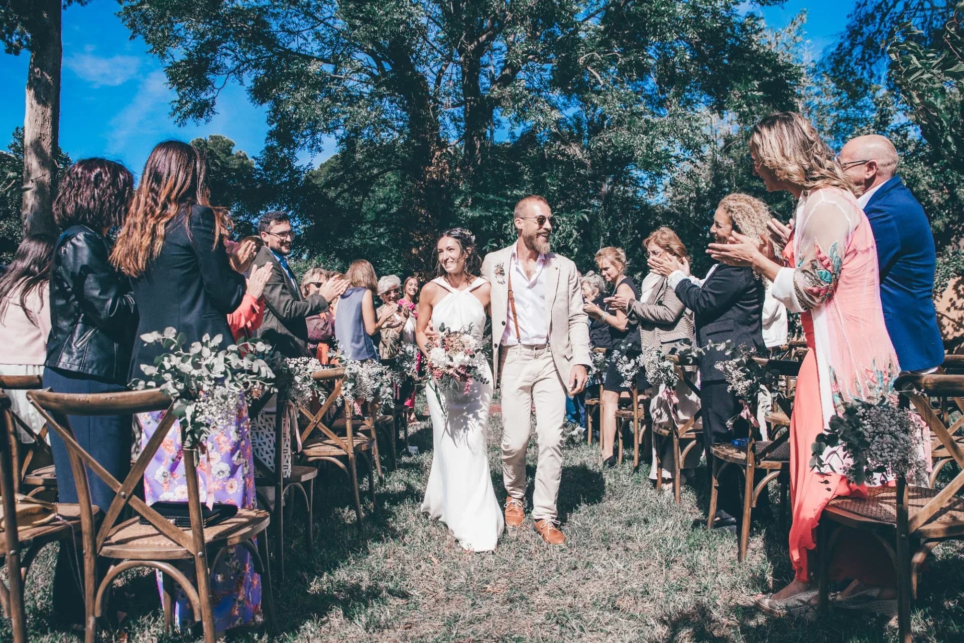 Los novios caminan tomados del brazo por el pasillo entre los invitados que aplauden sentados en sillas cruzadas con ramilletes de flores, bajo la sombra de grandes árboles en una ceremonia campestre.