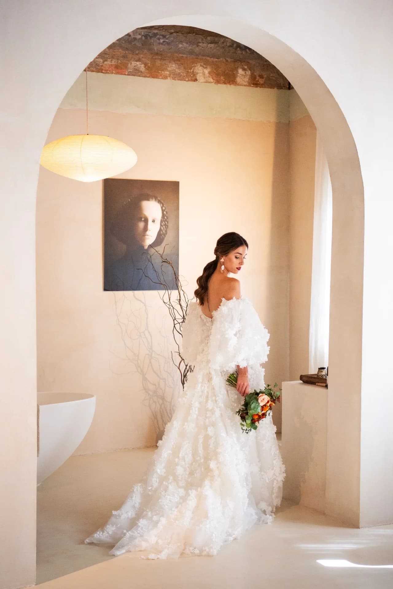 Novia de espaldas con vestido de encaje floral y mangas abullonadas, sosteniendo un ramo de flores, mirando hacia un cuadro colgado en una estancia de estilo mediterráneo con arco de obra vista y bañera exenta al fondo.