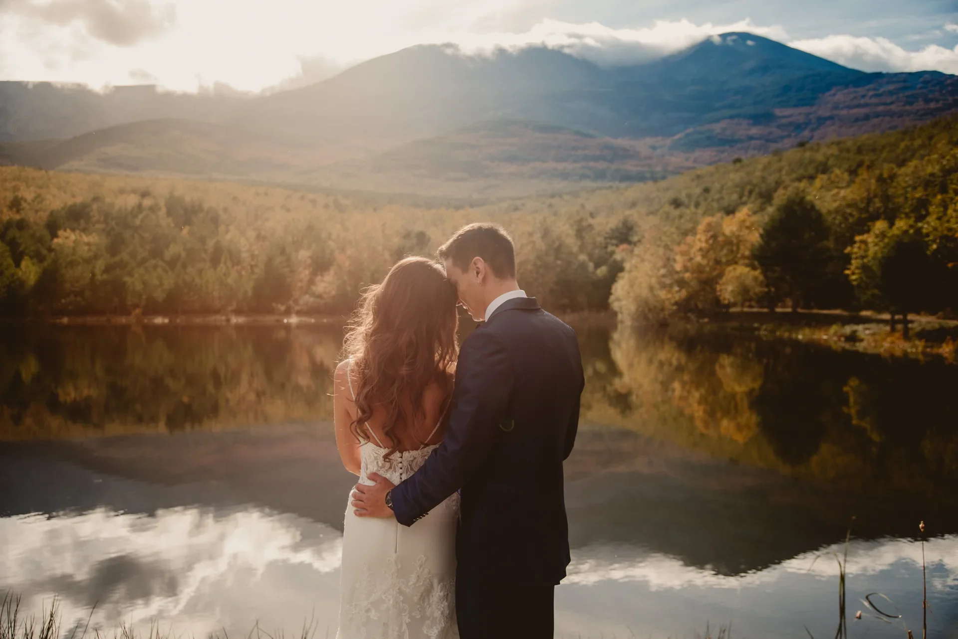 Pareja abrazada de espaldas frente a un lago en calma que refleja las montañas y el bosque otoñal, con el novio apoyando la frente sobre la cabeza de la novia mientras el sol entra a contraluz.