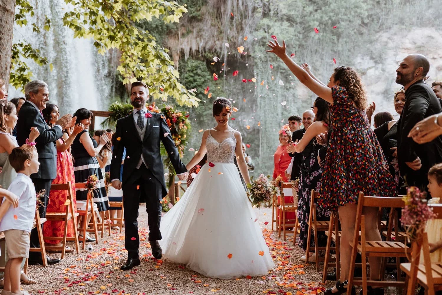 Los novios caminan sonrientes por el pasillo mientras los invitados les lanzan pétalos de flores, con una cascada y roca cubierta de musgo al fondo entre los árboles.