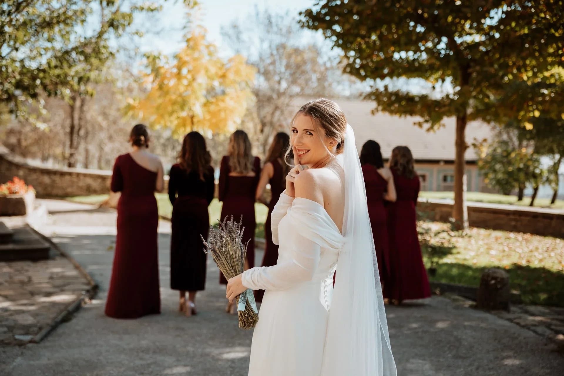 La novia mira hacia atrás sonriendo y con un dedo en los labios mientras sostiene un ramo de lavanda, con sus damas de honor vestidas de burdeos caminando de espaldas hacia un edificio entre árboles de otoño.
