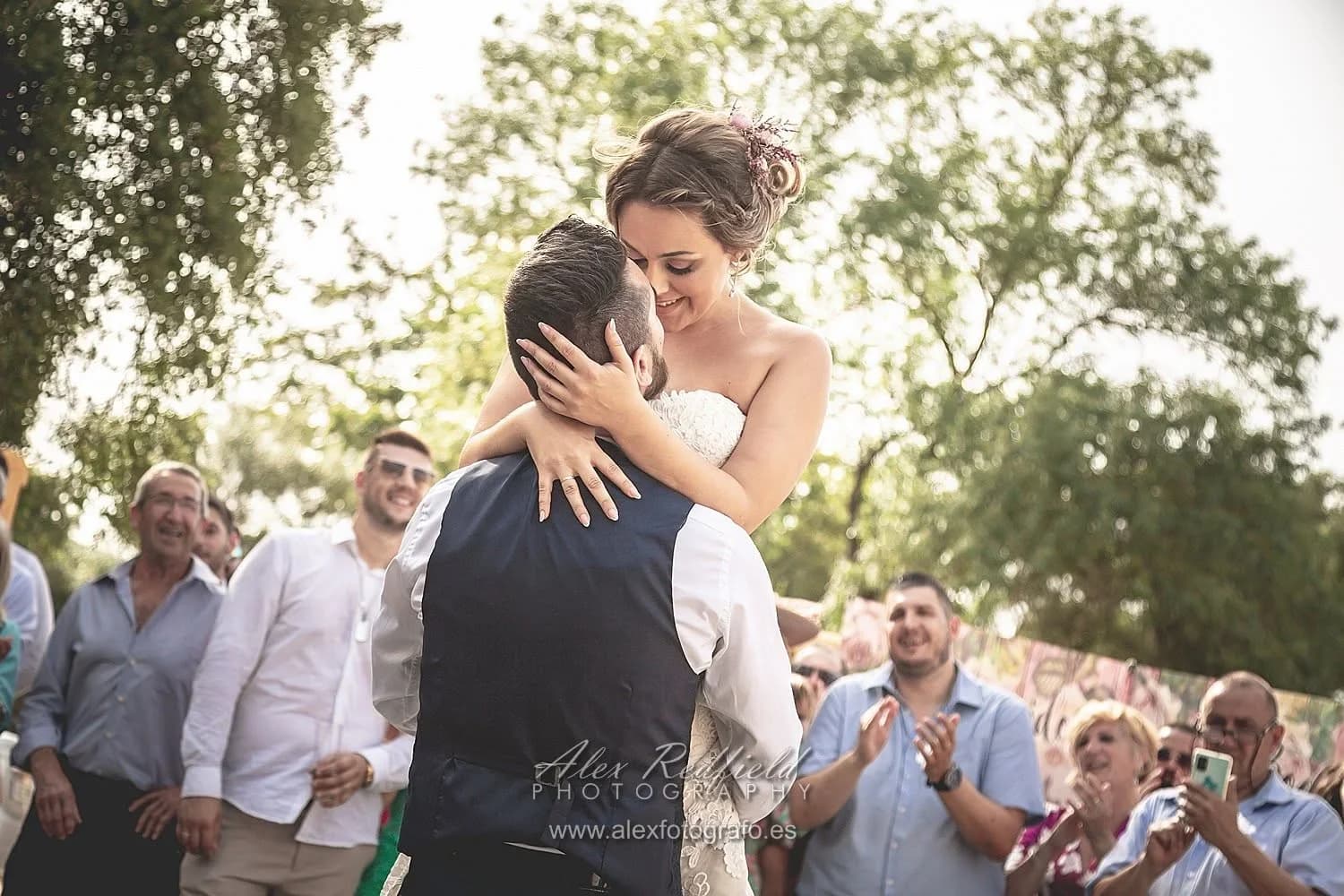 La novia es alzada en brazos por el novio durante el baile, rodeados de invitados que aplauden y sonríen entre árboles iluminados por el sol de la tarde.