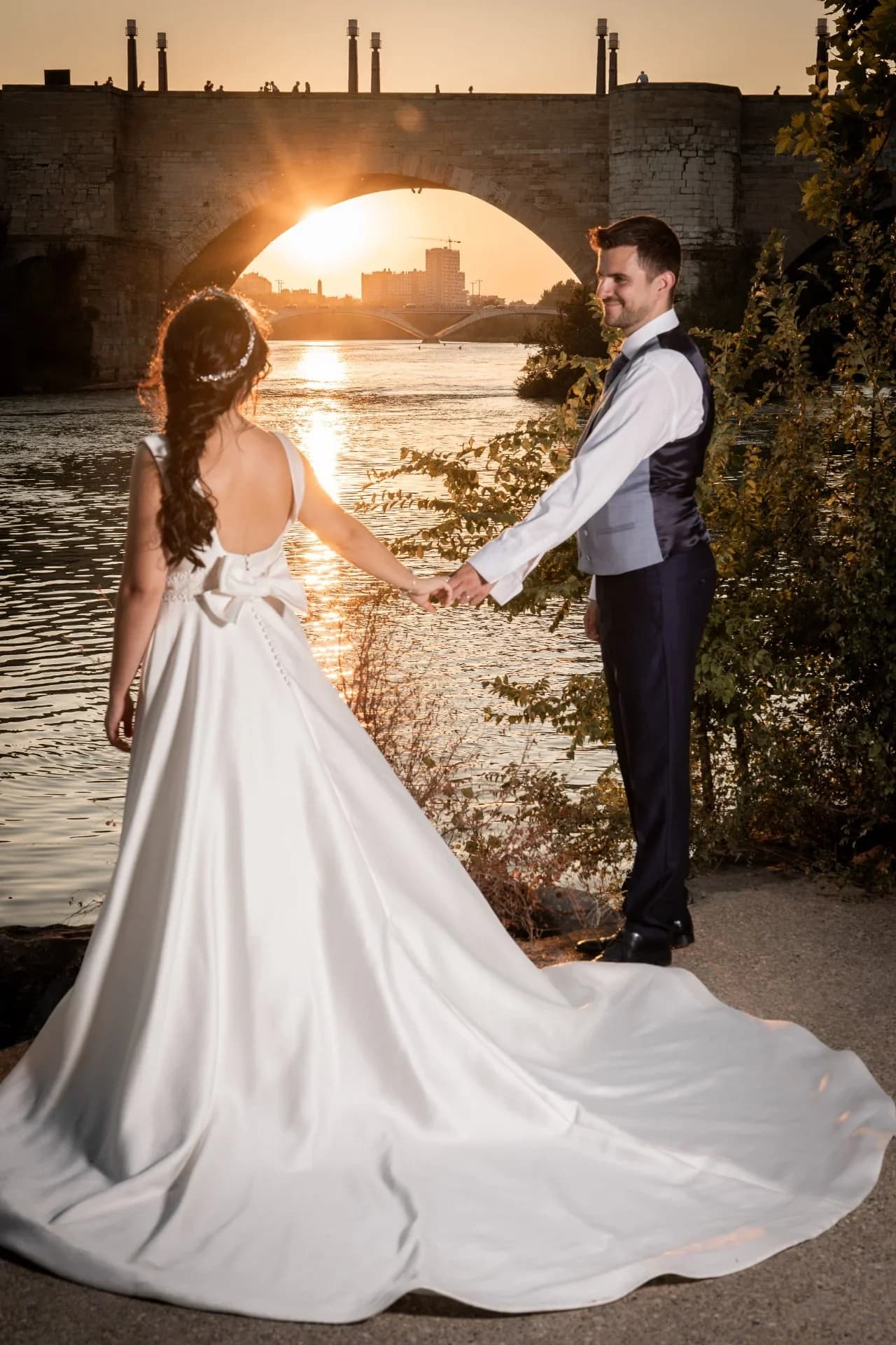 Los novios se dan la mano junto a un río al atardecer, vistos desde atrás de ella con la larga cola del vestido extendida sobre el suelo, mientras el sol se pone justo bajo el arco de un puente de piedra.