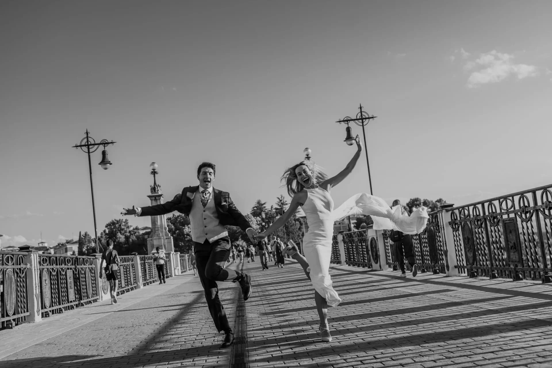 Pareja de recién casados corre saltando de la mano sobre un puente empedrado, en blanco y negro, con farolas antiguas de hierro forjado y transeúntes al fondo, mientras el vestido de ella vuela con el movimiento.