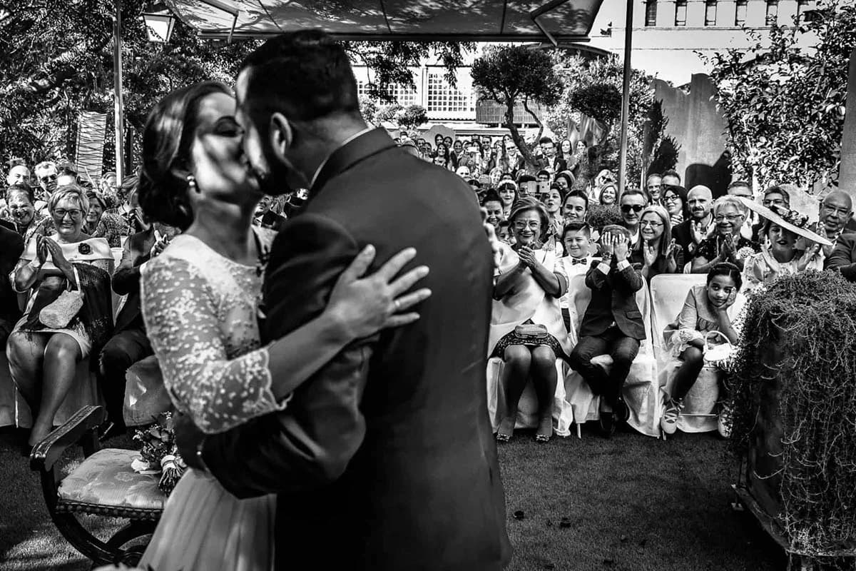 En blanco y negro, los novios se besan de espaldas al fotógrafo mientras los invitados sentados detrás aplauden y sonríen, en una ceremonia civil al aire libre bajo un toldo.