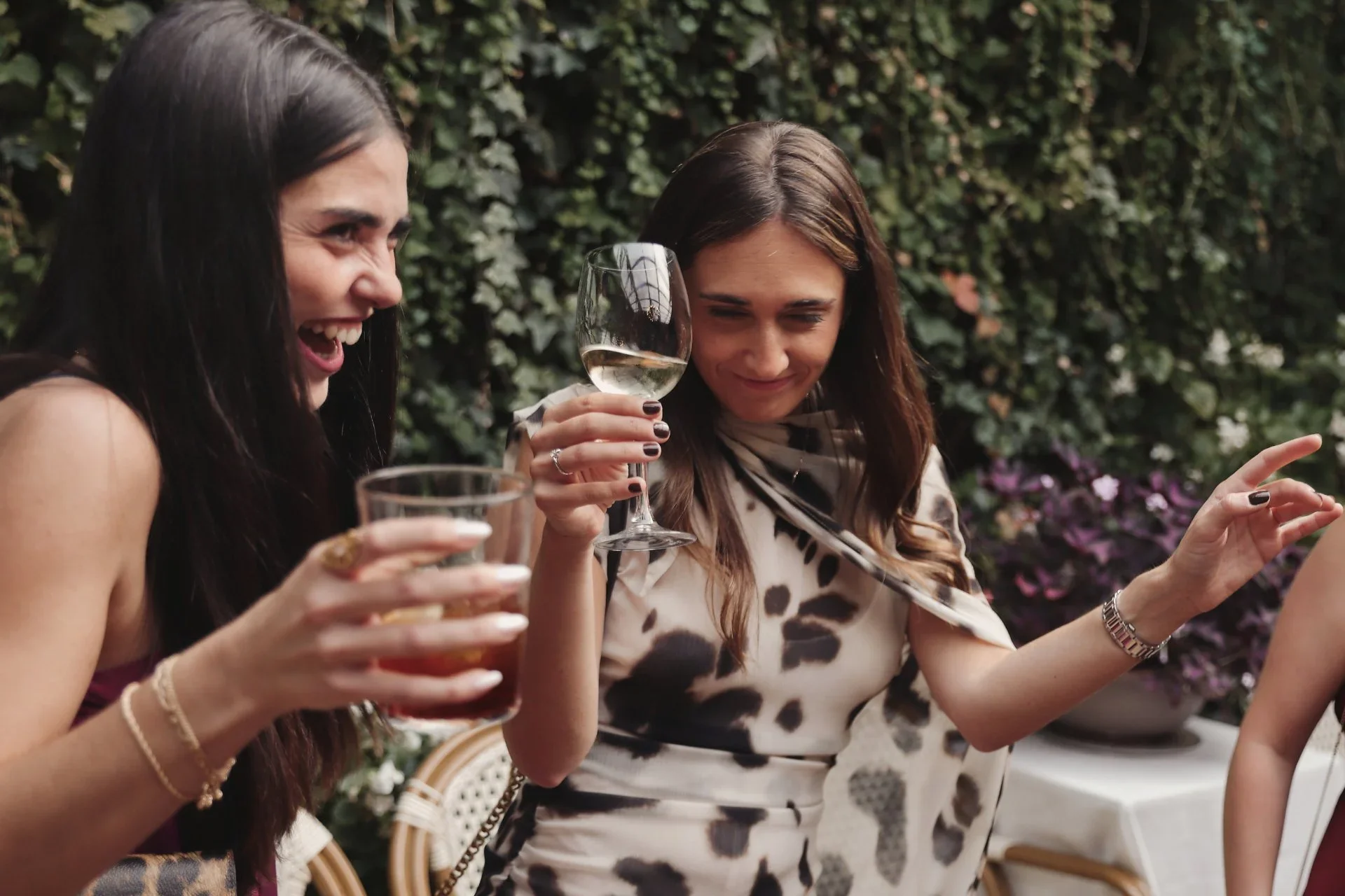 Dos invitadas ríen a carcajadas brindando con una copa de vino blanco y un cóctel, en un jardín con un muro cubierto de hiedra y flores moradas al fondo.