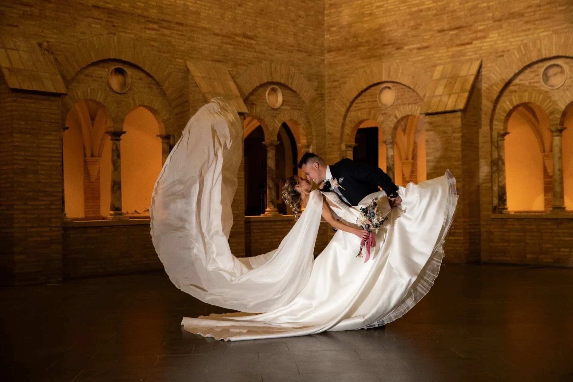 El novio inclina a la novia hacia atrás para besarla bajo unos arcos de ladrillo iluminados de tono cálido, mientras la larga cola de satén del vestido se despliega en el suelo formando una curva envolvente.