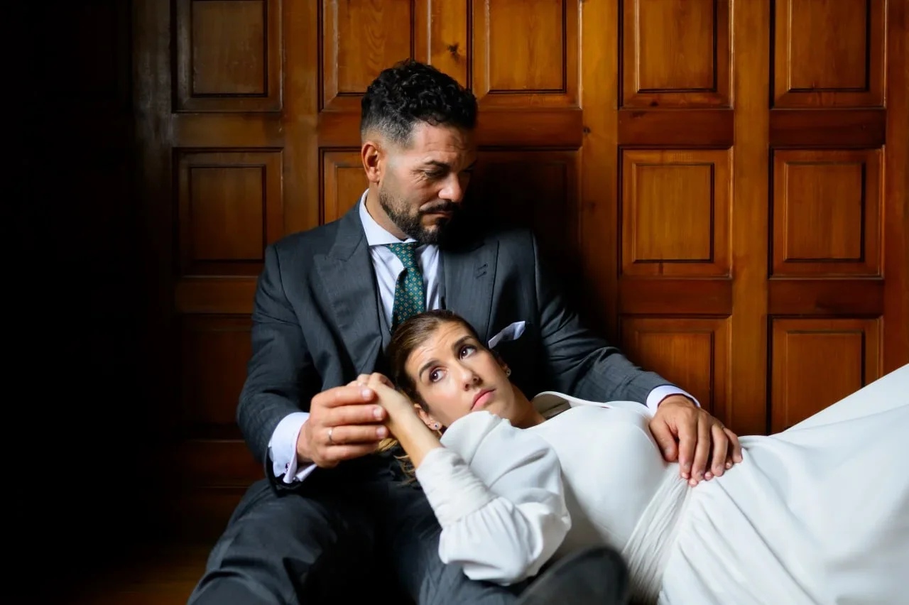 Los novios sentados en el suelo apoyados contra una puerta de madera tallada, ella recostada con la cabeza sobre el regazo y las manos de él mientras lo mira de reojo, en un momento íntimo y relajado tras la ceremonia.