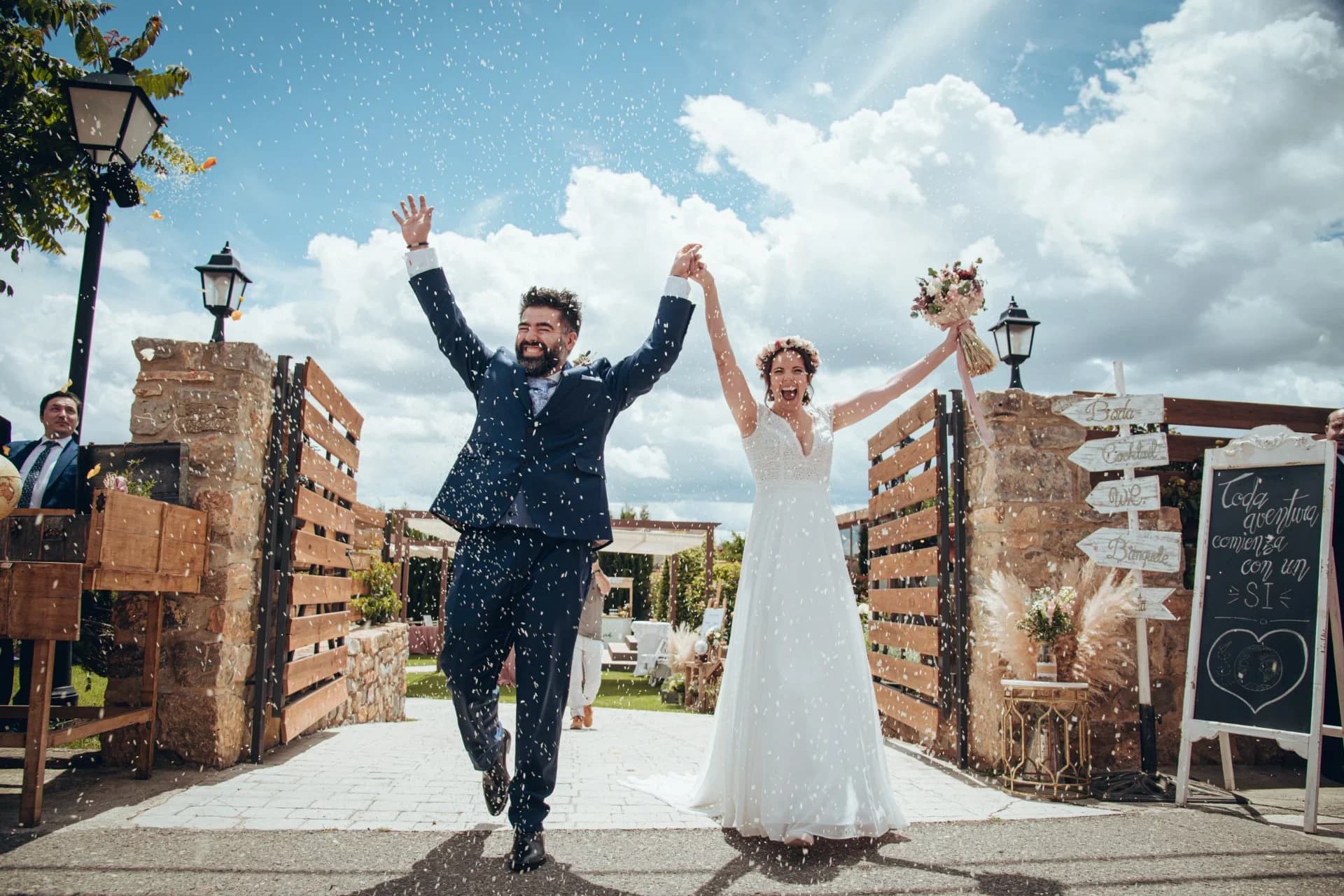 El novio y la novia corren tomados de la mano y riendo bajo una lluvia de arroz, junto a un cartel de madera que dice 'Toda aventura comienza con un sí' y un cielo azul con nubes.