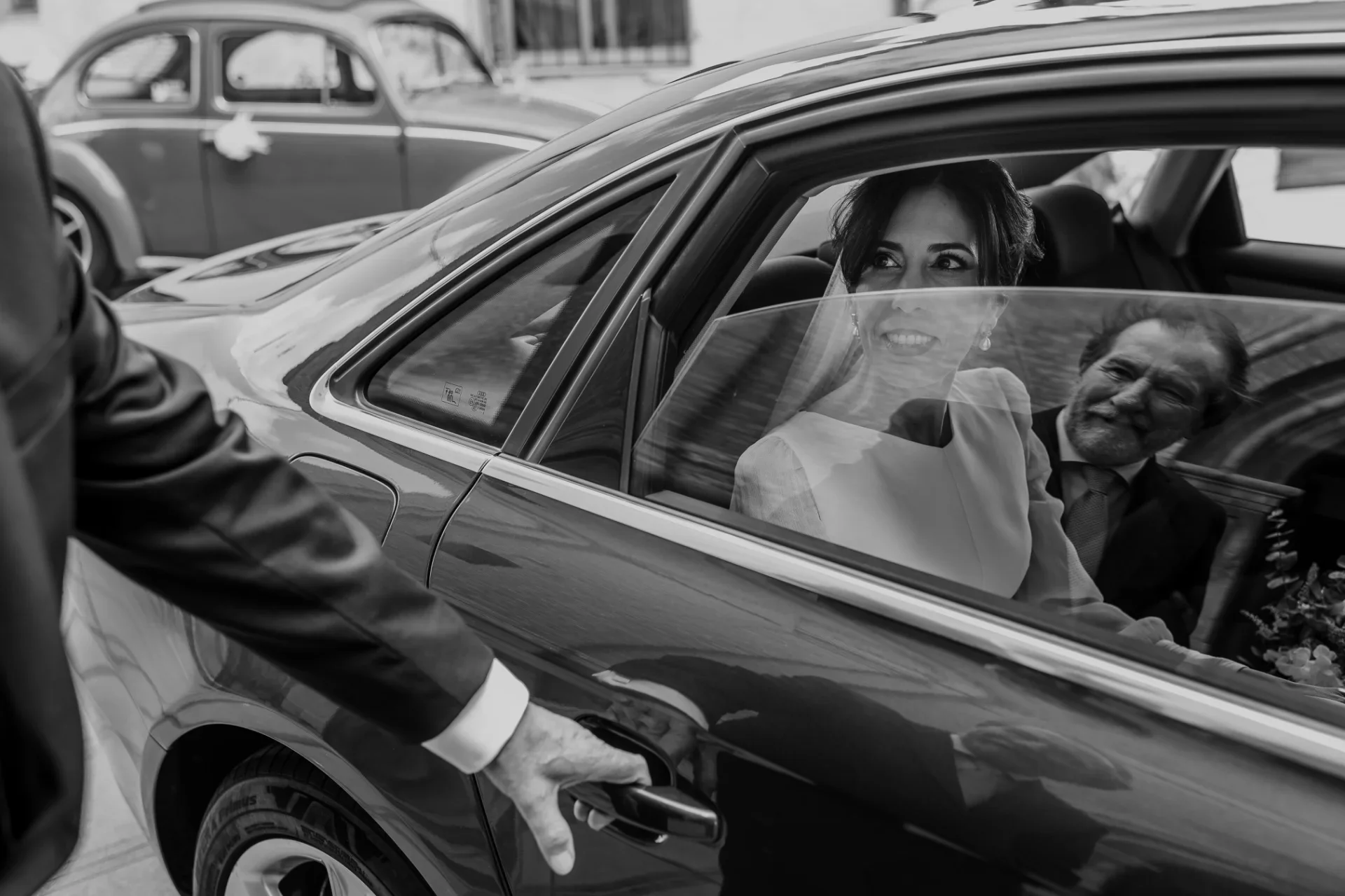 En blanco y negro, la novia sonríe desde el asiento trasero de un coche clásico mientras un hombre le abre la puerta desde fuera; se aprecia el reflejo de otro coche de época al fondo de la calle.