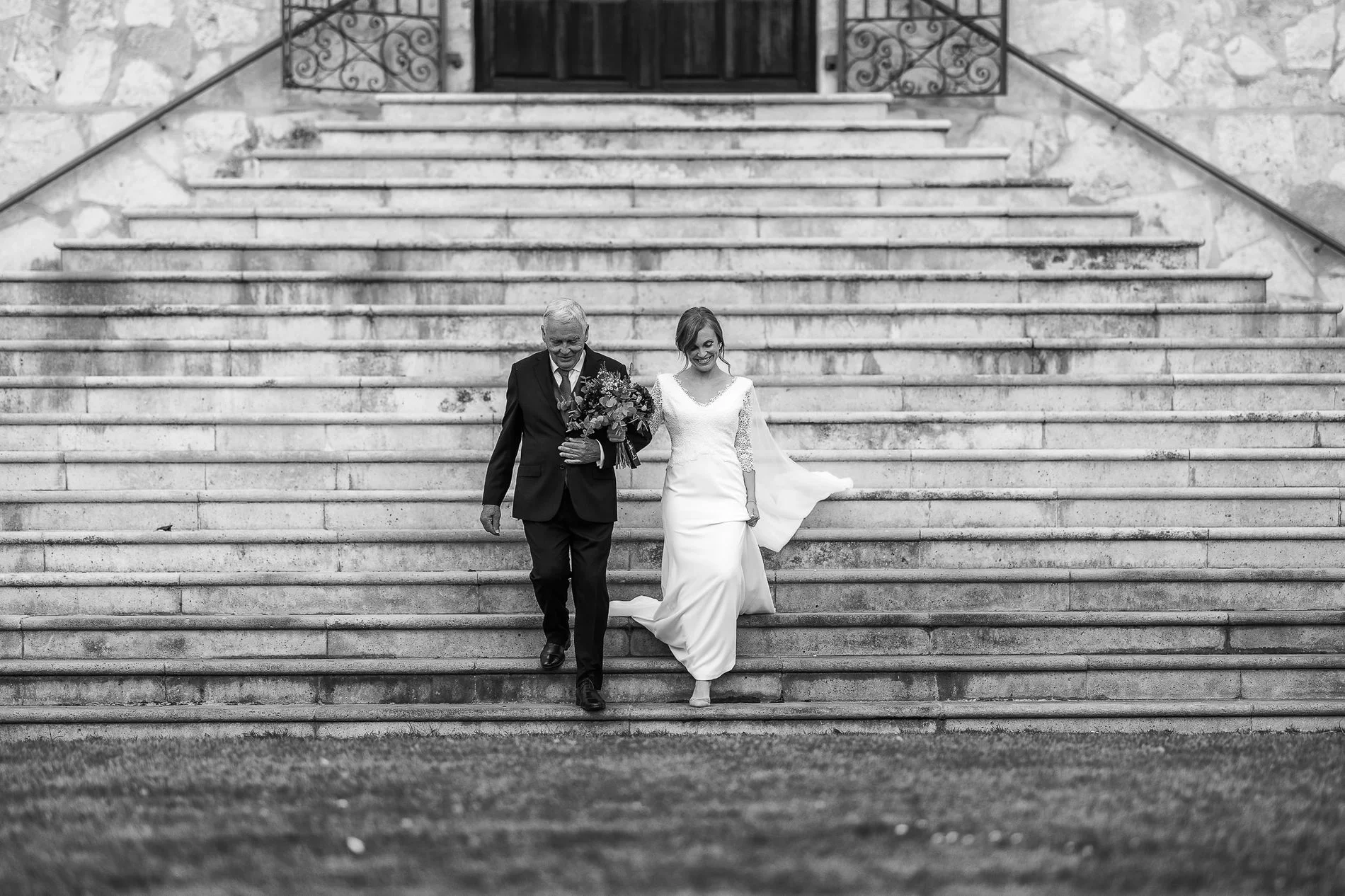 Fotografía en blanco y negro del padre de la novia acompañándola mientras bajan juntos una gran escalinata de piedra frente a una puerta de hierro forjado, ella riendo con un ramo de flores oscuras.