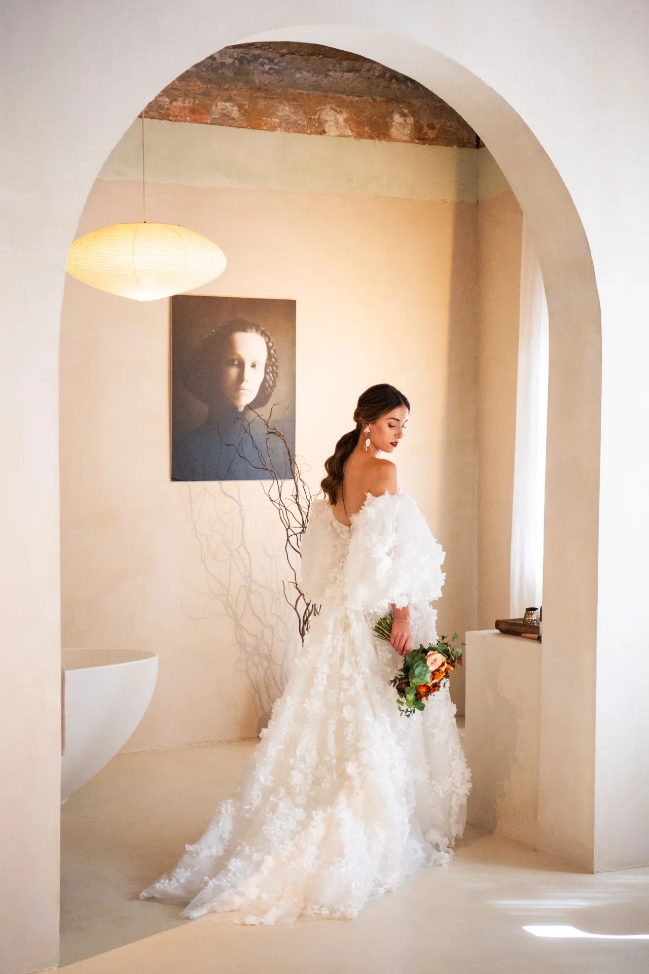 Novia de espaldas con vestido de encaje floral y mangas abullonadas, sosteniendo un ramo de flores, mirando hacia un cuadro colgado en una estancia de estilo mediterráneo con arco de obra vista y bañera exenta al fondo.
