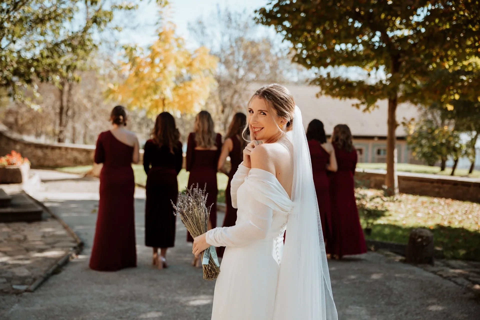 La novia mira hacia atrás sonriendo y con un dedo en los labios mientras sostiene un ramo de lavanda, con sus damas de honor vestidas de burdeos caminando de espaldas hacia un edificio entre árboles de otoño.
