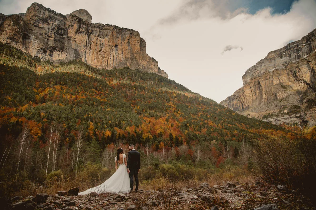 Los novios de espaldas, cogidos de la mano, contemplan un valle de montaña con paredes rocosas y laderas cubiertas de bosque otoñal en tonos ocre y rojo bajo un cielo nublado.