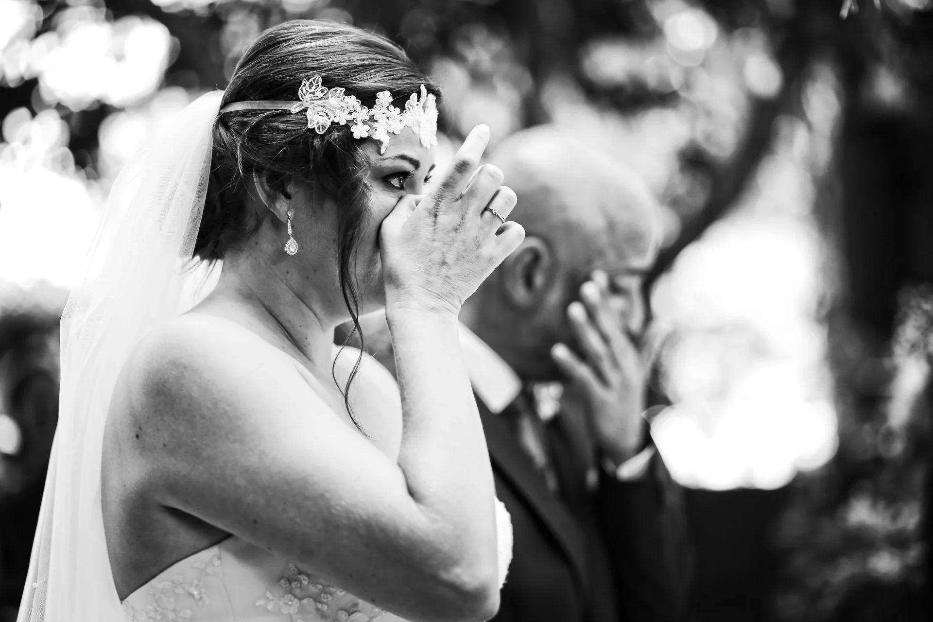 Fotografía en blanco y negro de una novia emocionada llevándose la mano a la boca mientras llora durante la ceremonia, con un hombre desenfocado aplaudiendo detrás de ella.