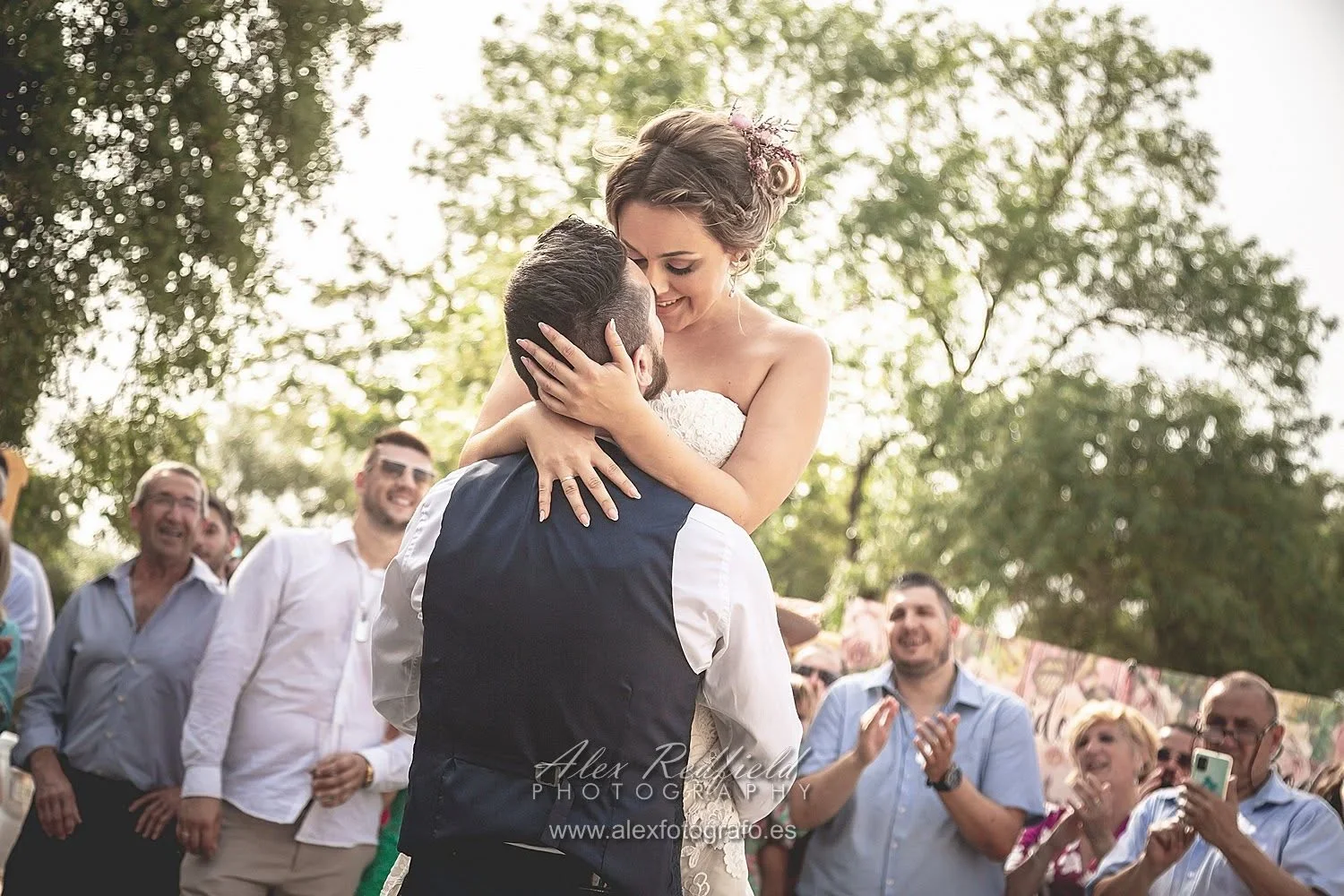 La novia es alzada en brazos por el novio durante el baile, rodeados de invitados que aplauden y sonríen entre árboles iluminados por el sol de la tarde.