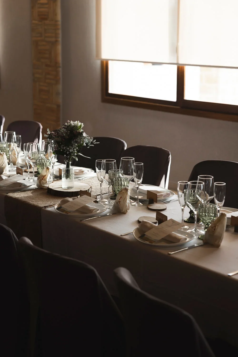 Mesa larga de banquete vista en perspectiva diagonal, con manteles claros, copas de cristal transparente y verde, servilletas de tela anudadas con el menú y un pequeño centro floral silvestre junto a una rodaja de madera, iluminada por luz de ventana lateral.