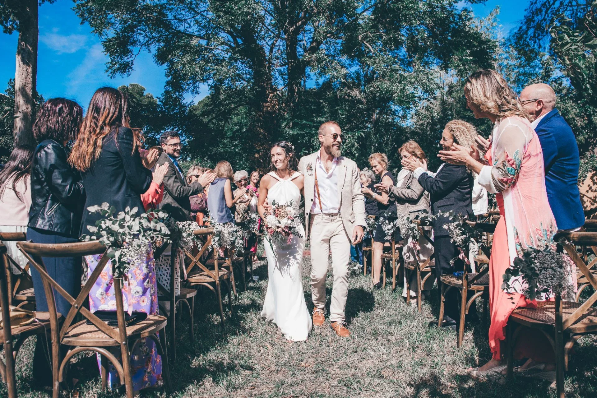 Los novios caminan tomados del brazo por el pasillo entre los invitados que aplauden sentados en sillas cruzadas con ramilletes de flores, bajo la sombra de grandes árboles en una ceremonia campestre.