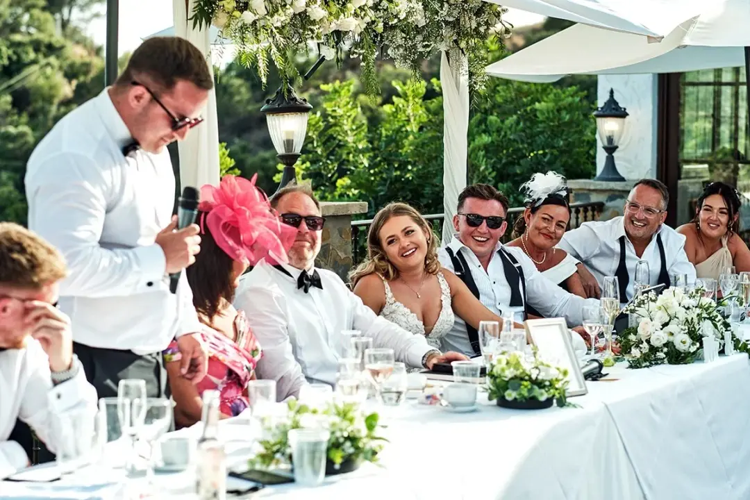 Mesa presidencial en una terraza al aire libre durante un banquete de boda, un hombre de pie con micrófono da un discurso mientras la novia y otros invitados, algunos con gafas de sol y tocados, ríen a carcajadas bajo un arco floral.