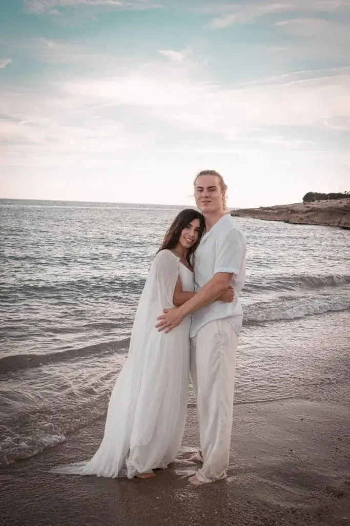 Pareja abrazada de pie en la orilla del mar, vestidos de blanco, mirando a cámara con el horizonte marino y una cala rocosa al fondo, cielo nublado suave.