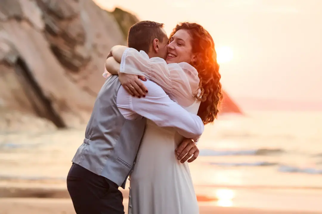 Pareja abrazándose y riendo en una playa con acantilados al atardecer, ella con el pelo rizado pelirrojo y él con chaleco gris, el sol reflejándose sobre el mar en calma.