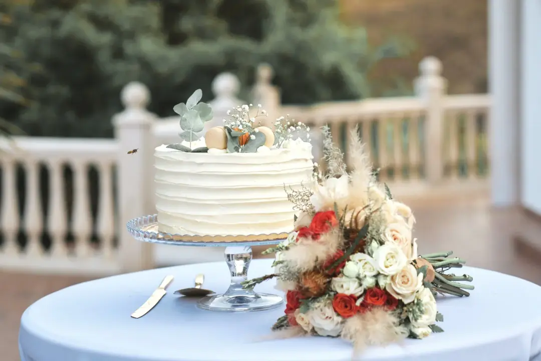 Detalle de una tarta nupcial de un piso decorada con macarons, eucalipto y flores blancas junto a un ramo de novia de rosas rojas y blancas con pampas, sobre una mesa con mantel azul claro y balaustrada desenfocada al fondo.