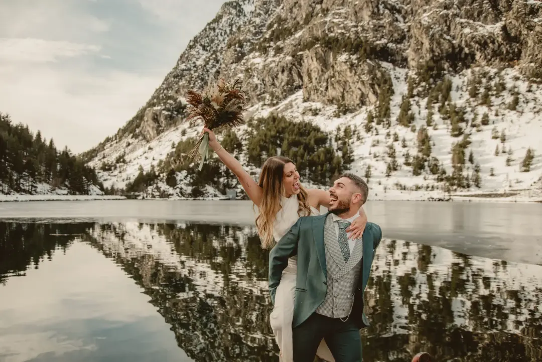 El novio lleva a la novia a caballito junto a un lago de montaña nevado, ella riendo y levantando un ramo de flores secas hacia el cielo, con picos rocosos y nieve al fondo.