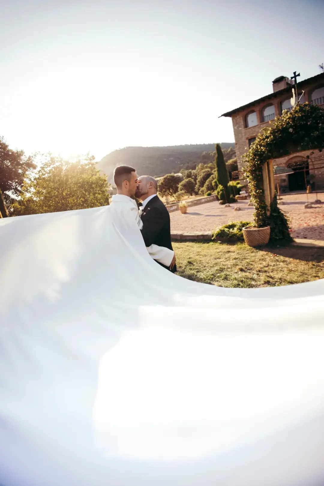 Dos novios se besan al atardecer frente a una masía catalana con arco de piedra y buganvilla, mientras la larga capa blanca de uno de ellos ondea en primer plano cubriendo casi toda la imagen.
