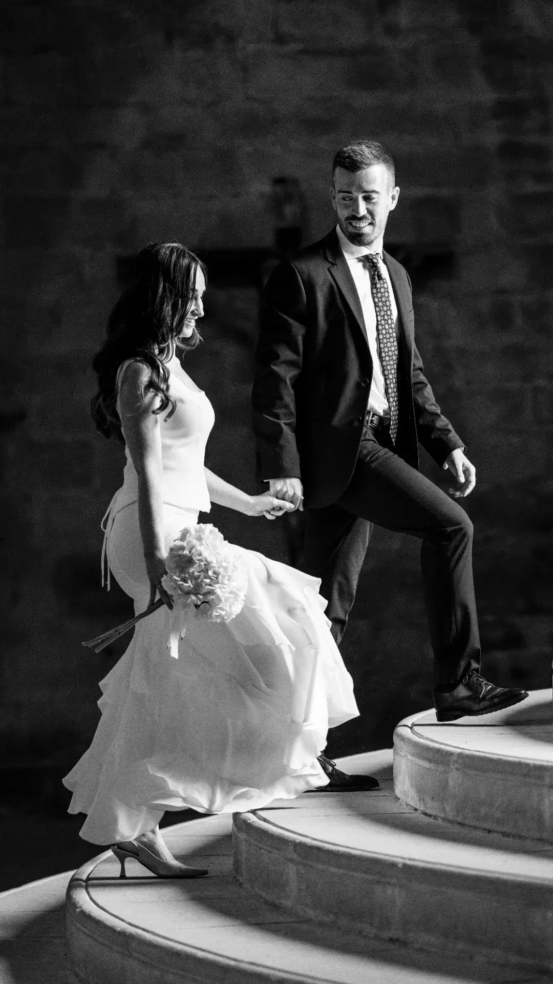 El novio levanta a la novia en volandas durante el baile nocturno bajo palmeras y guirnaldas de luces, ella con los brazos extendidos hacia el cielo, mientras invitados aplauden y graban con el móvil al fondo.