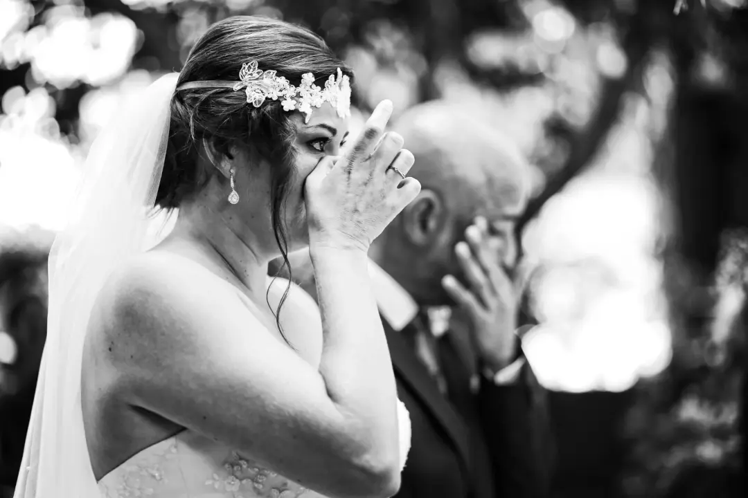 Fotografía en blanco y negro de una novia emocionada llevándose la mano a la boca mientras llora durante la ceremonia, con un hombre desenfocado aplaudiendo detrás de ella.