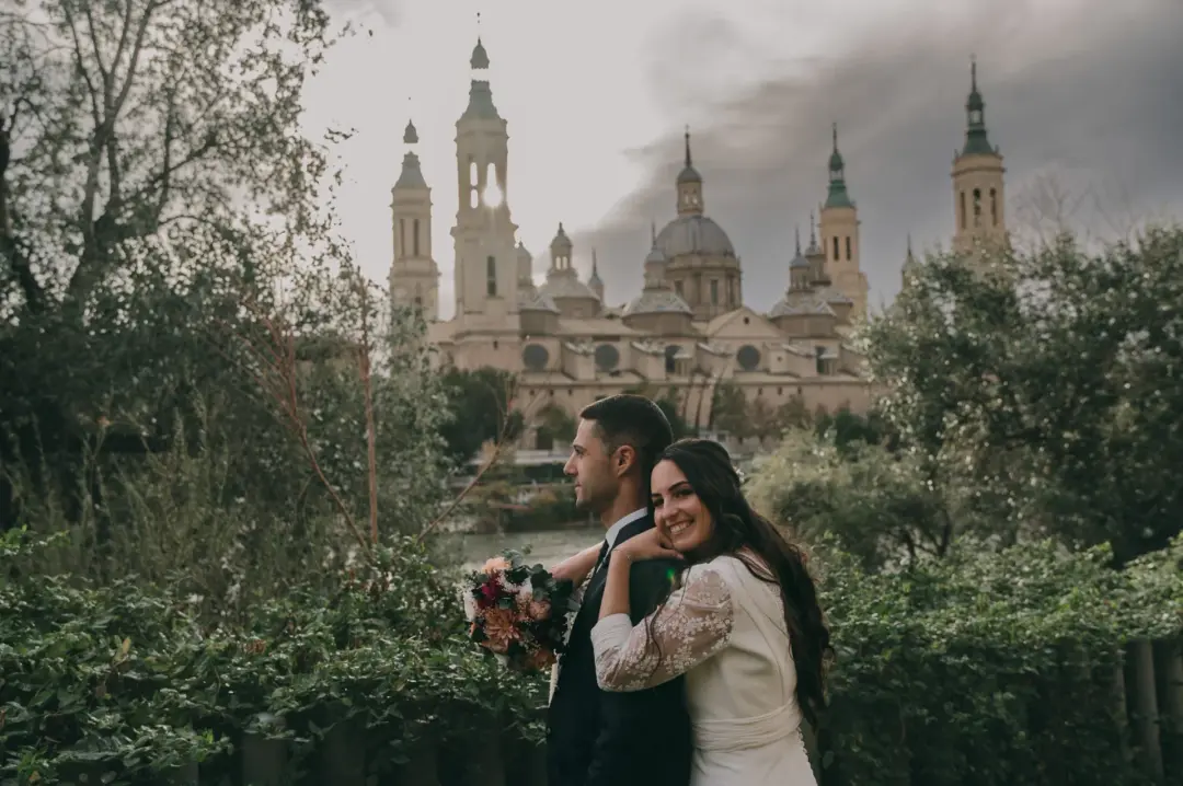 Pareja de novios abrazada, ella sonriendo hacia la cámara con un ramo de flores en tonos coral y burdeos, frente a la Basílica del Pilar de Zaragoza bajo un cielo nublado y dramático, rodeados de vegetación.