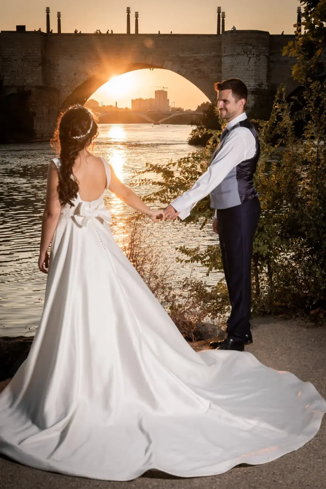 Los novios se dan la mano junto a un río al atardecer, vistos desde atrás de ella con la larga cola del vestido extendida sobre el suelo, mientras el sol se pone justo bajo el arco de un puente de piedra.